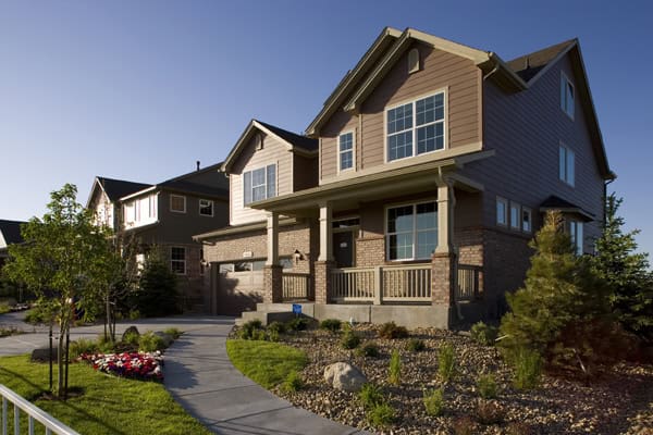 Two-story suburban house sits illuminated by late-afternoon sun, showing a covered porch and large windows; landscaped yard with curved concrete walkway, flower beds, rocks and neighboring homes in the background.