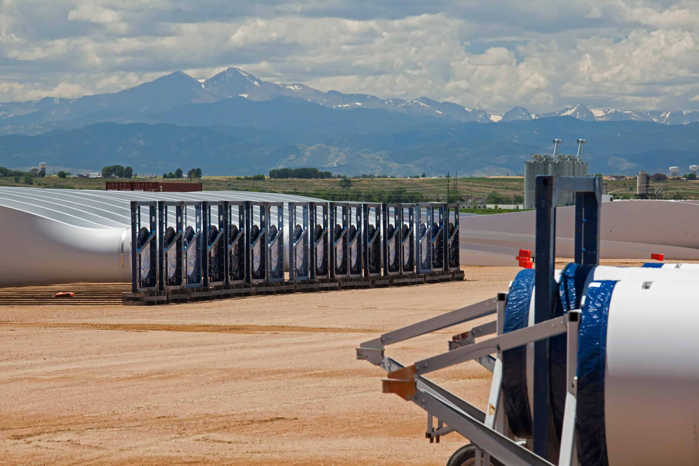 Rows of white wind-turbine blades lie stacked in metal racks, stored on a dusty construction yard with industrial tanks and distant snow-capped mountains under a cloudy sky.