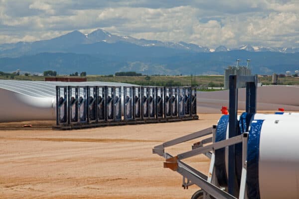 Rows of white wind-turbine blades lie stacked in metal racks, stored on a dusty construction yard with industrial tanks and distant snow-capped mountains under a cloudy sky.