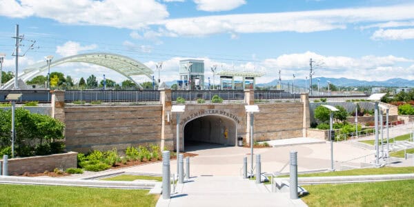 Stone arch reading "WESTMINSTER STATION" opens to a tunnel amid a landscaped transit plaza under an elevated light‑rail platform and white arched bridge, with distant mountains and blue sky.