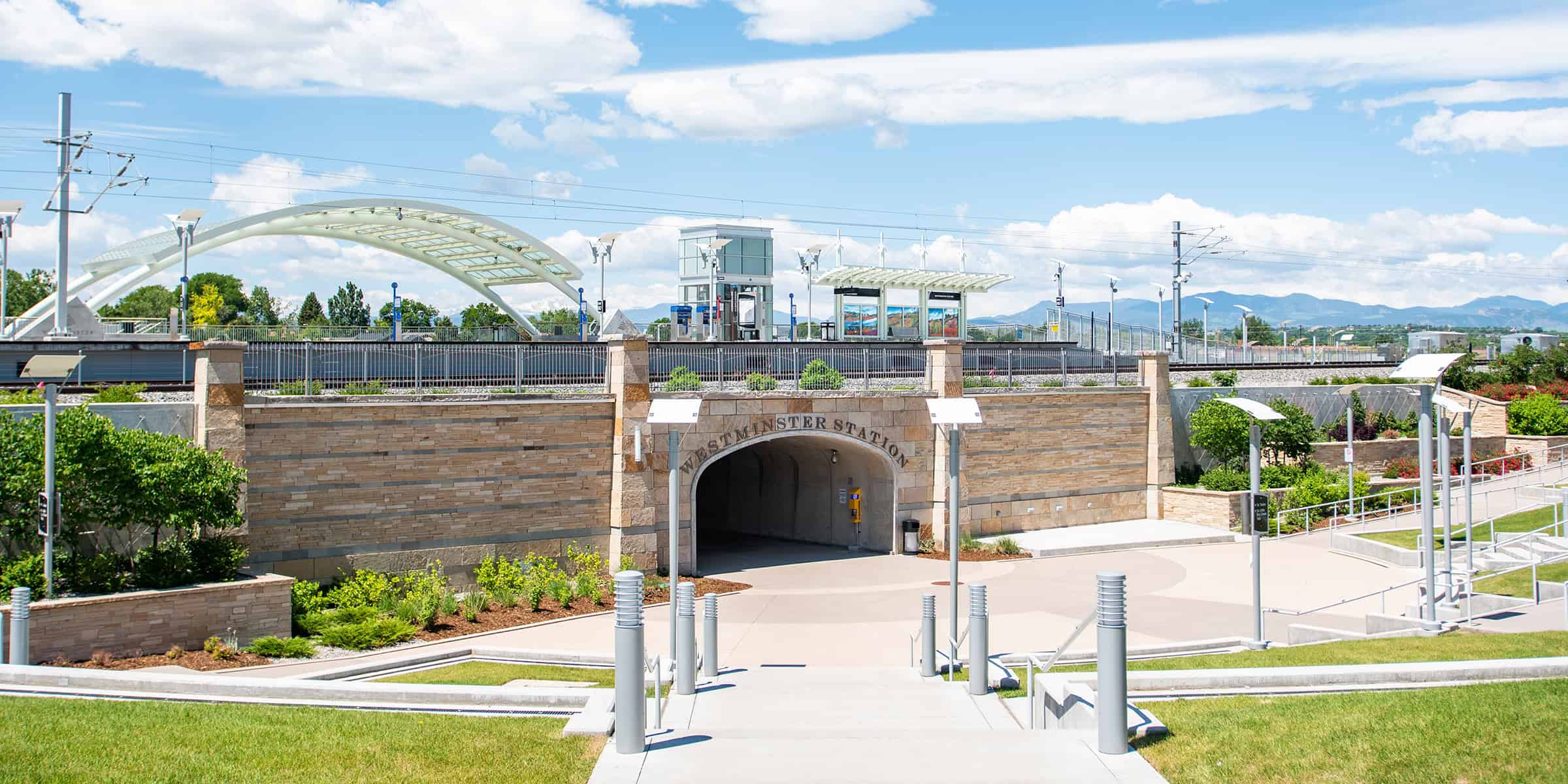 Stone arch reading "WESTMINSTER STATION" opens to a tunnel amid a landscaped transit plaza under an elevated light‑rail platform and white arched bridge, with distant mountains and blue sky.