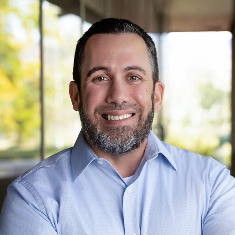 Bearded man smiling with arms crossed, wearing a light-blue dress shirt; standing indoors by large windows with soft natural light and blurred green trees visible outside.