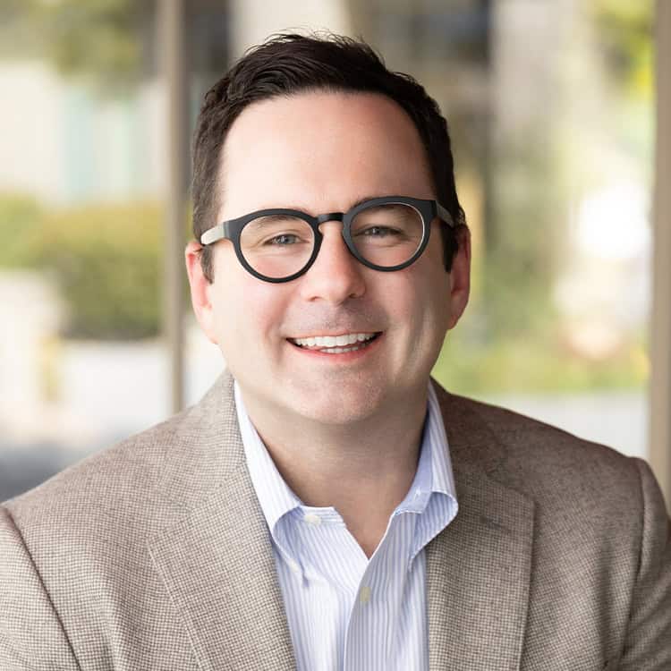Man wearing round black glasses smiles toward the camera, dressed in a brown tweed blazer and light-blue striped shirt, seated before a softly blurred outdoor/office background.