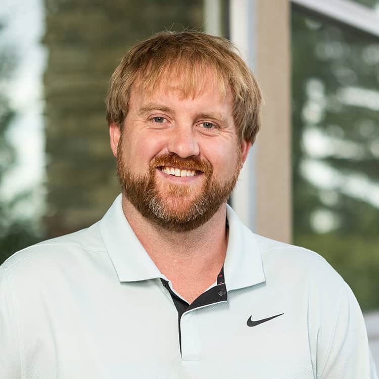 A smiling bearded adult man with light brown hair faces the camera, wearing a light polo with a swoosh logo; indoors near a window overlooking blurred trees.