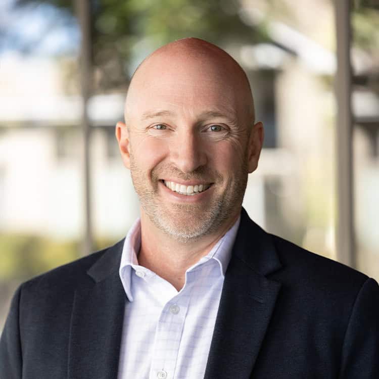 Bald, smiling middle-aged man looking at the camera in a softly blurred modern interior with large windows and greenery visible outside.