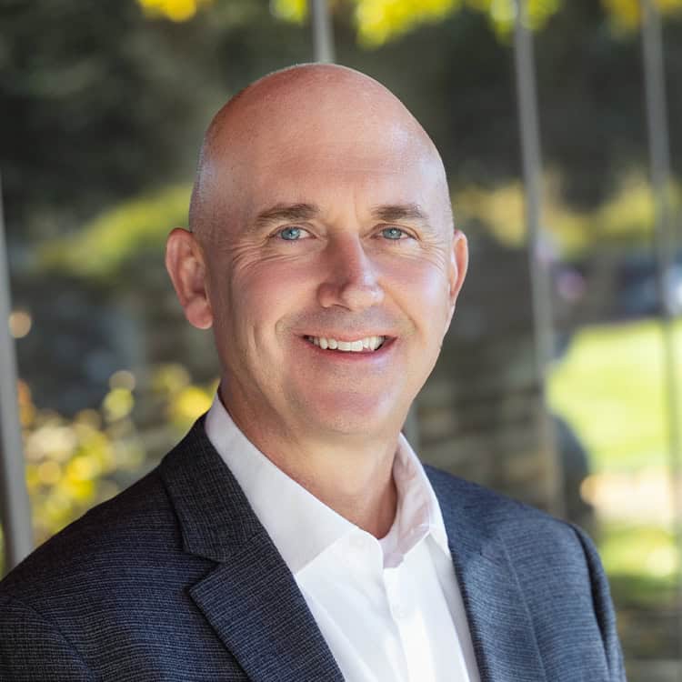 A bald, middle-aged man smiles at the camera, wearing a white shirt and dark blazer, posed outdoors in front of glass windows with blurred green foliage reflected behind him.
