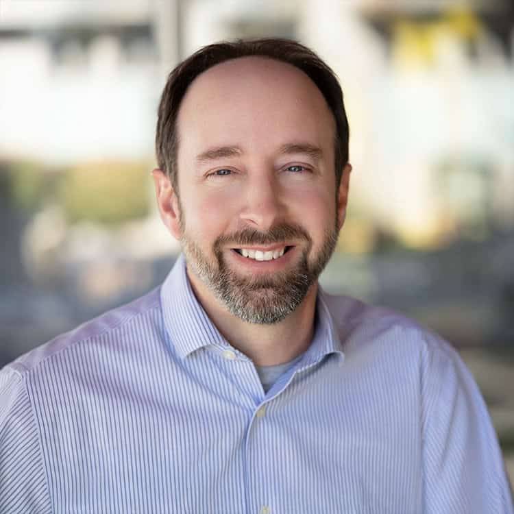 Bearded man smiles directly at the camera, wearing a light-blue striped button-down shirt; soft, blurred sunlit background suggests an indoor workspace or café.