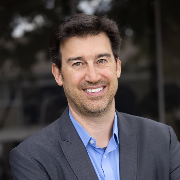 Smiling middle-aged man in a gray blazer and blue shirt poses facing the camera, standing outdoors before a blurred glass building reflection.