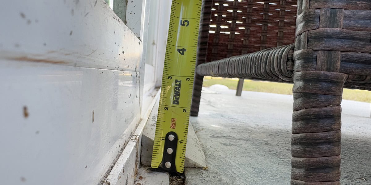 A yellow tape measure stands vertically, measuring a small gap at a door track while a wicker chair leg sits nearby on a concrete patio.

Text in image: DEWALT, 25ft, 1 2 3 4 5