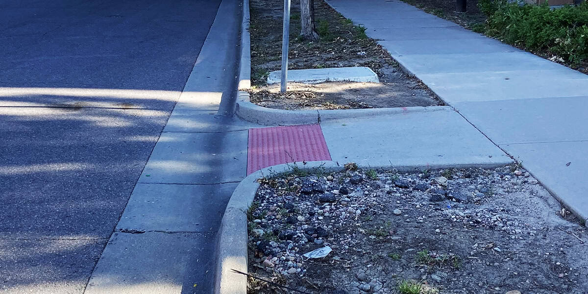 Curb ramp with red tactile paving slopes down to an asphalt street, connecting a concrete sidewalk; adjacent rocky dirt patch, signpost, and tree shadows line the residential curb.