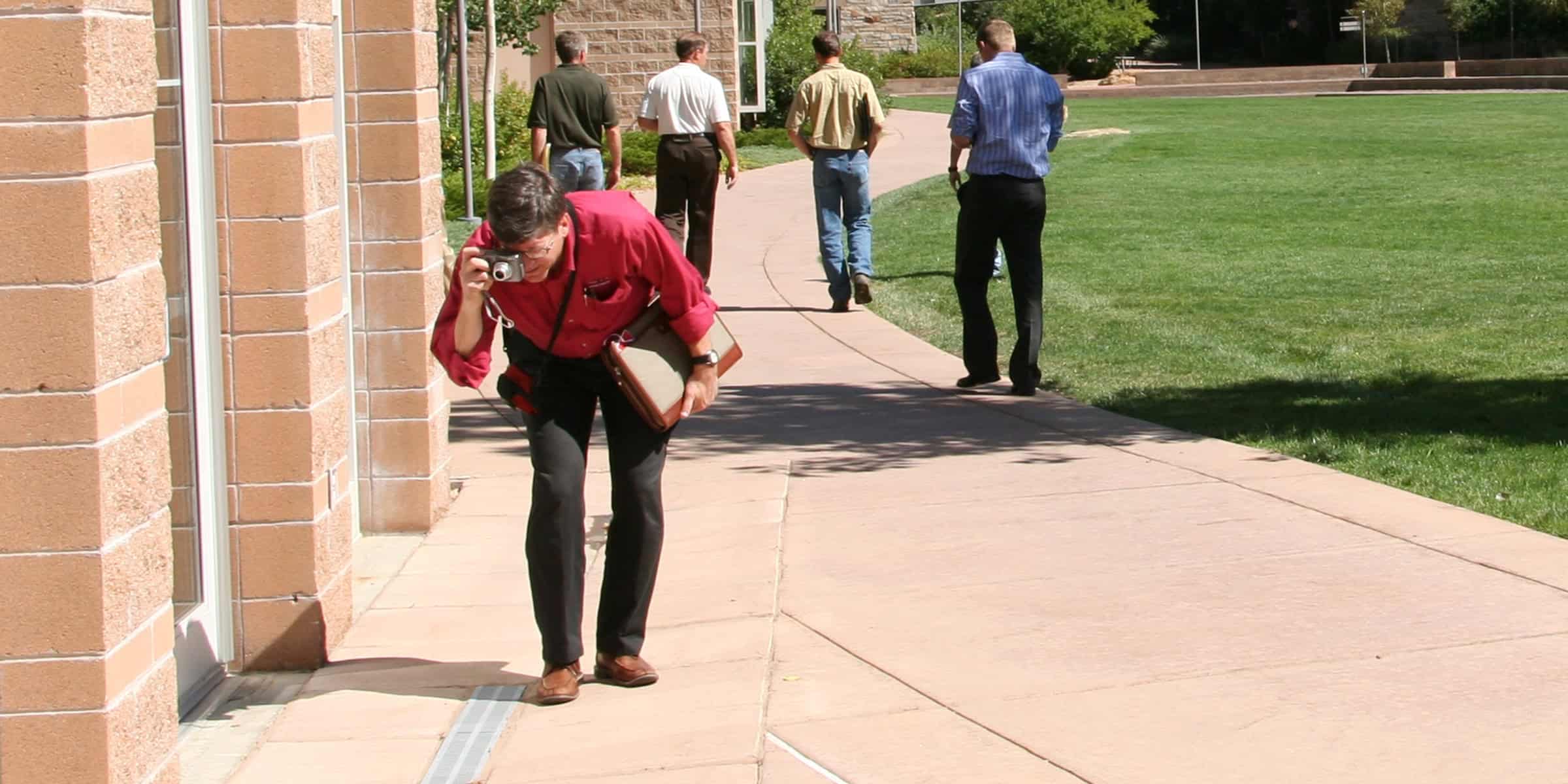 Man in red shirt leans forward, photographing with a compact camera while holding a briefcase on a sidewalk beside a brick building; several men walk away across a nearby lawn.