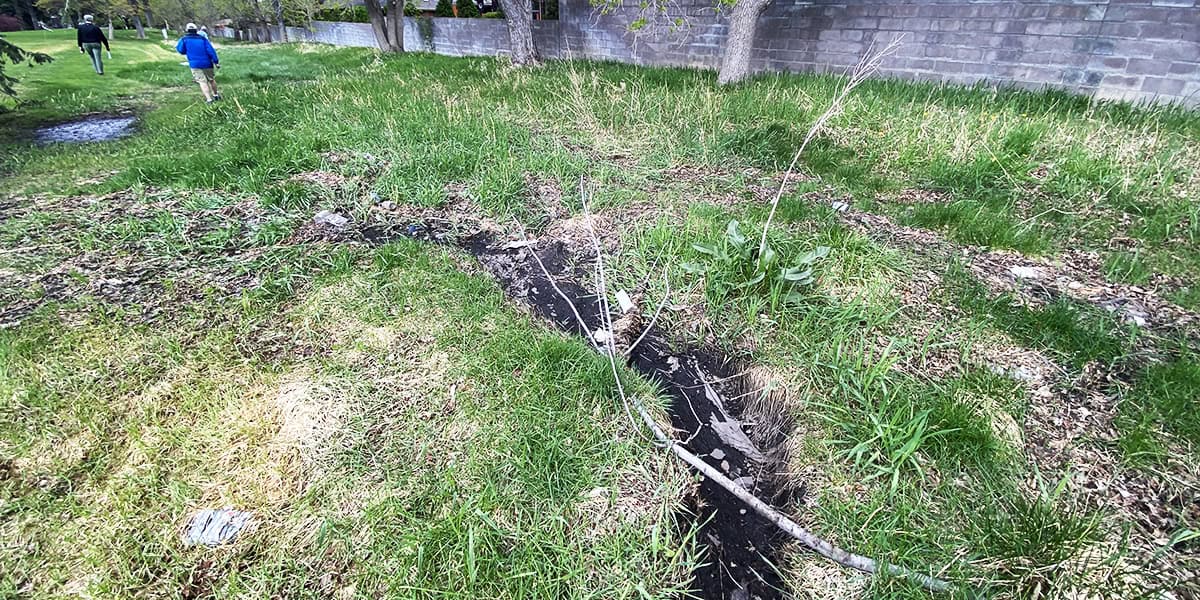 A narrow eroded soil gully cuts through overgrown grass; a fallen branch lies across it. Two people walk away near trees and a concrete-block wall in the background.