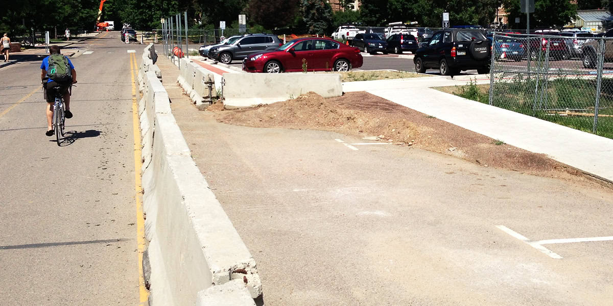 Concrete barrier runs down street, separating a blocked bike lane from traffic; a cyclist rides past on the open lane, with parked cars, chain-link fence, and trees in the background.