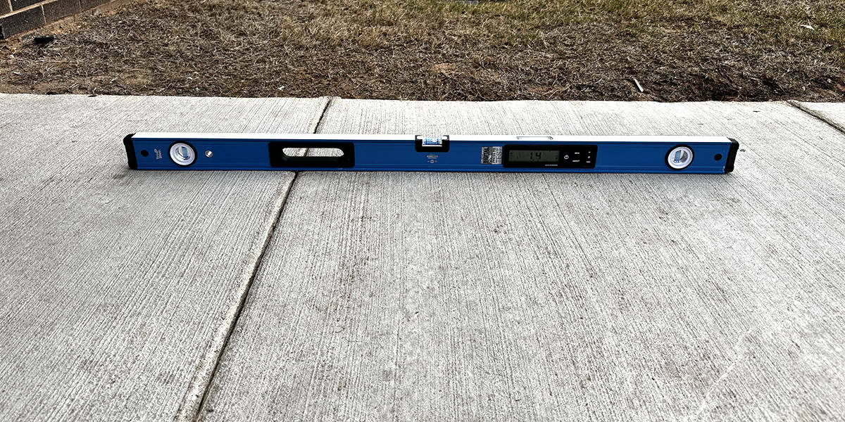 Blue digital spirit level lies across a sidewalk seam, showing a 1.4° tilt on its display; concrete slabs in the foreground, dried grass and soil border the top edge.