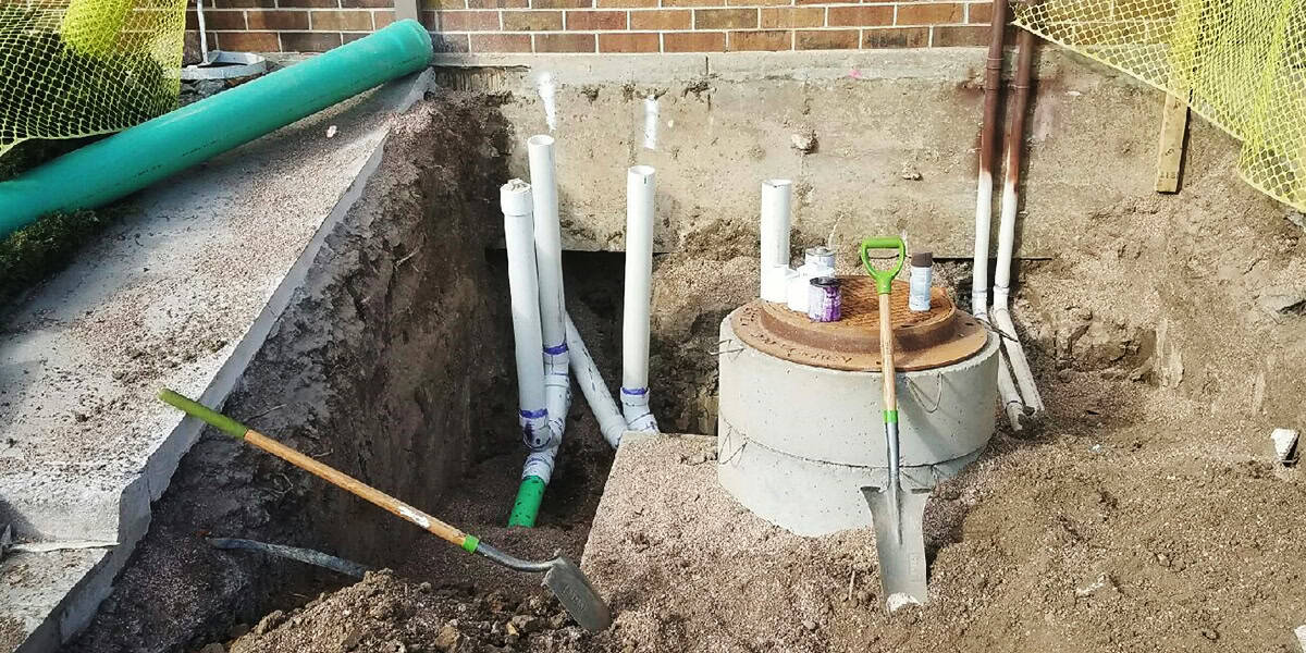 Round concrete riser with rusted lid sits in a dug trench; vertical PVC drain pipes and a green pipe nearby, two shovels leaning in loose soil before a brick wall.