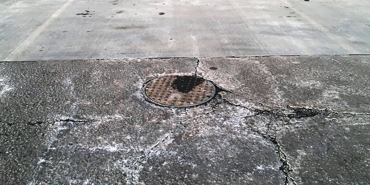 Round metal manhole cover sits embedded in cracked concrete, surrounded by radiating fissures and white salt stains, set in an empty, weathered concrete lot with a smoother slab beyond.