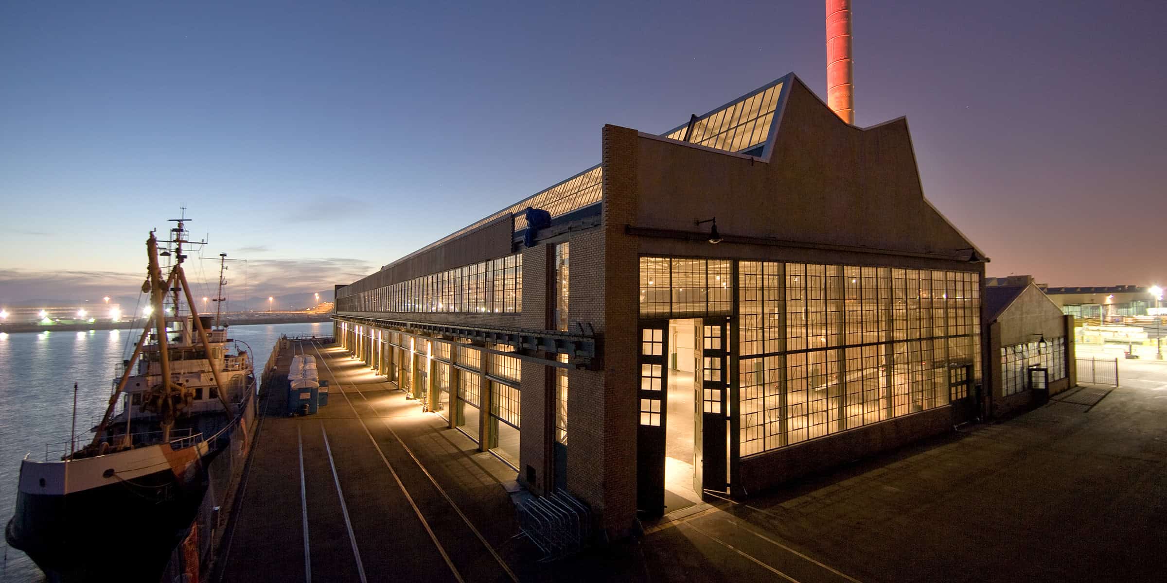 Industrial waterfront warehouse glows through large grid windows, its open doorway and interior lights illuminating a quay where a moored ship rests alongside under twilight sky and distant harbor lights.