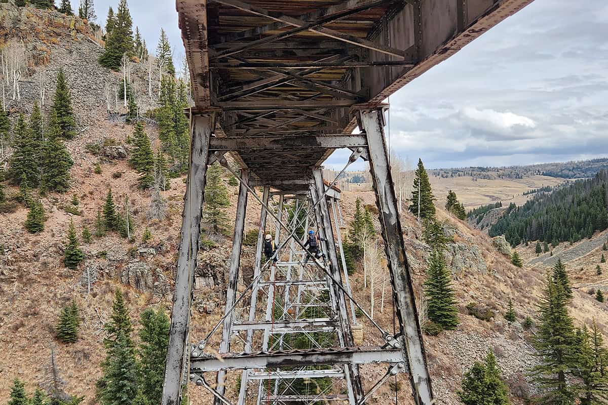 A rusted steel railroad trestle spans a canyon; two helmeted, harnessed climbers traverse its central truss above steep, pine- and rock-covered slopes beneath a cloudy sky.