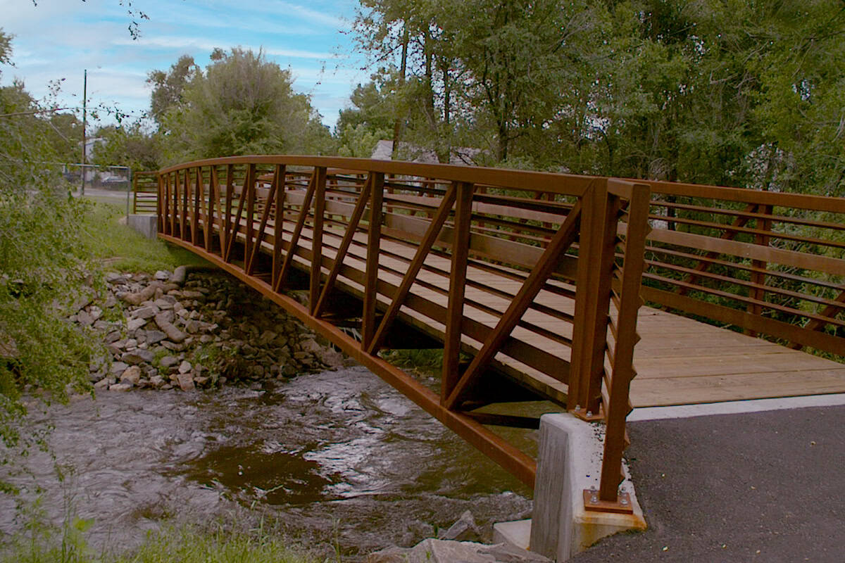 A rust-colored metal footbridge spans a shallow rocky stream, its wooden planks and triangular rail supports crossing between concrete abutments amid trees and grassy banks under a blue sky.