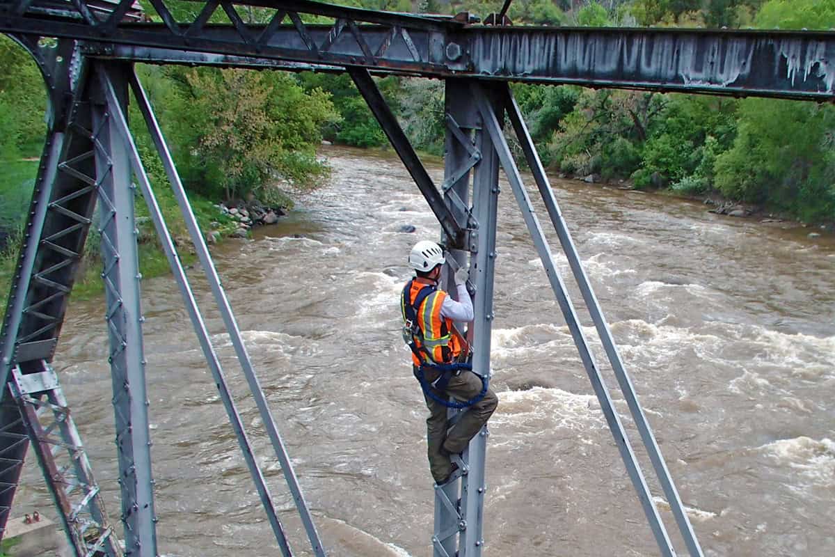 Bridge worker in a safety harness and helmet climbs a vertical steel support, inspecting bolts, above a fast, muddy river framed by bridge trusses and green riverside trees.