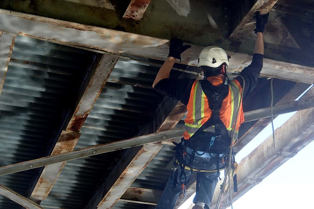 A construction worker in a white helmet and orange safety vest reaches upward to inspect or repair rusted steel girders beneath a corrugated metal bridge, secured by a safety harness.