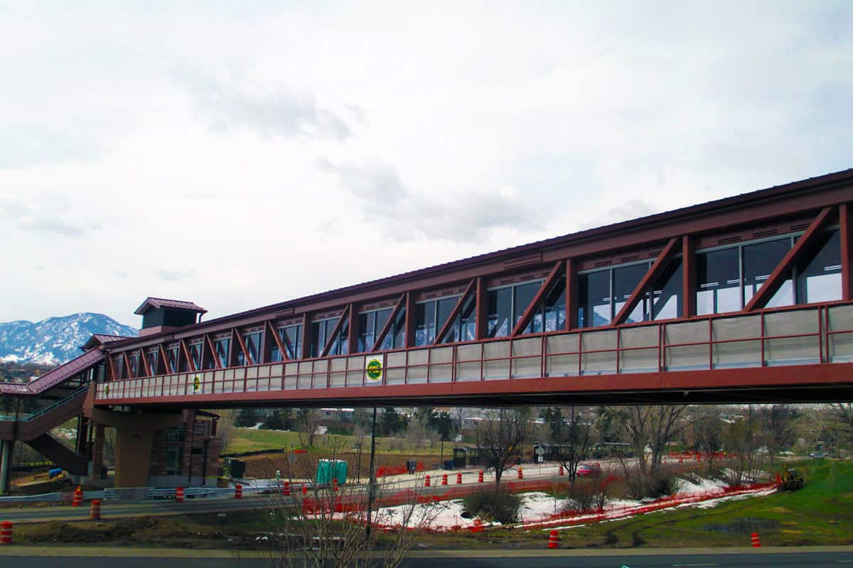 Enclosed pedestrian skybridge spans a roadway, connecting stairs at left; construction barrels and orange fencing surround a partly snow-covered grassy median, with mountains and cloudy sky beyond.