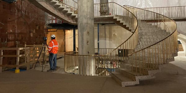 Spiral concrete staircase rises around a central column with brass railings; a worker in an orange high-visibility jacket operates a tripod-mounted surveying instrument nearby, inside a dim, unfinished construction interior.
