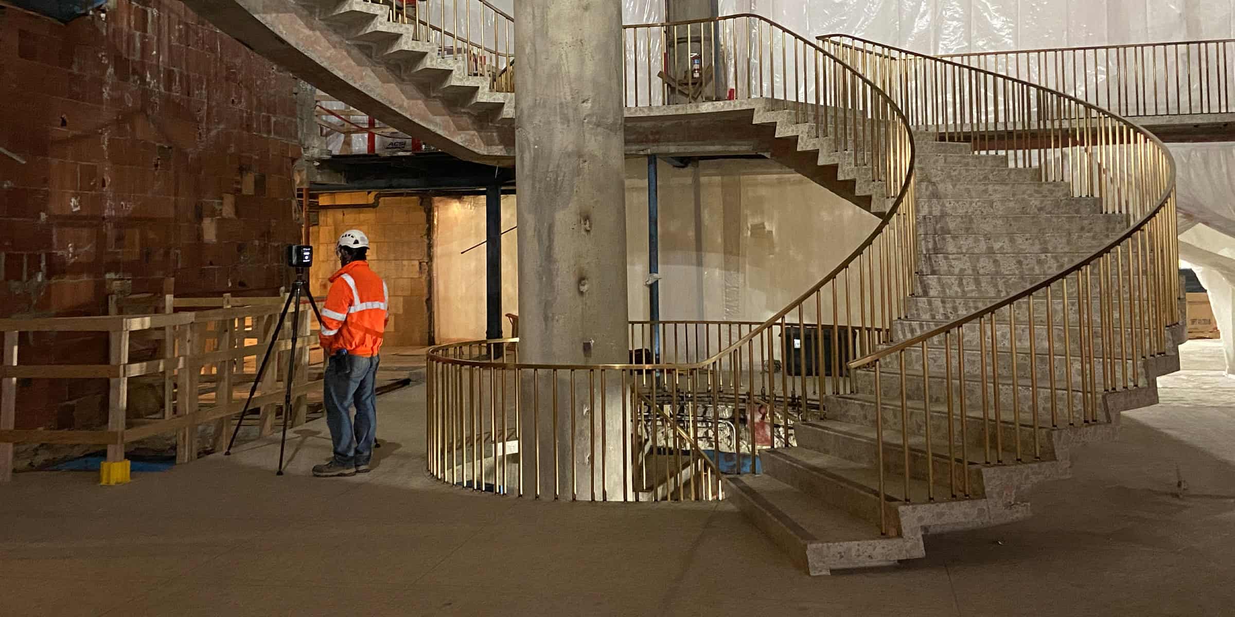 Spiral concrete staircase rises around a central column with brass railings; a worker in an orange high-visibility jacket operates a tripod-mounted surveying instrument nearby, inside a dim, unfinished construction interior.