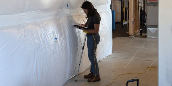 Woman using a tablet mounted on a surveying pole, tapping the screen while standing beside large plastic-wrapped insulation in a construction basement with exposed framing and concrete floor.