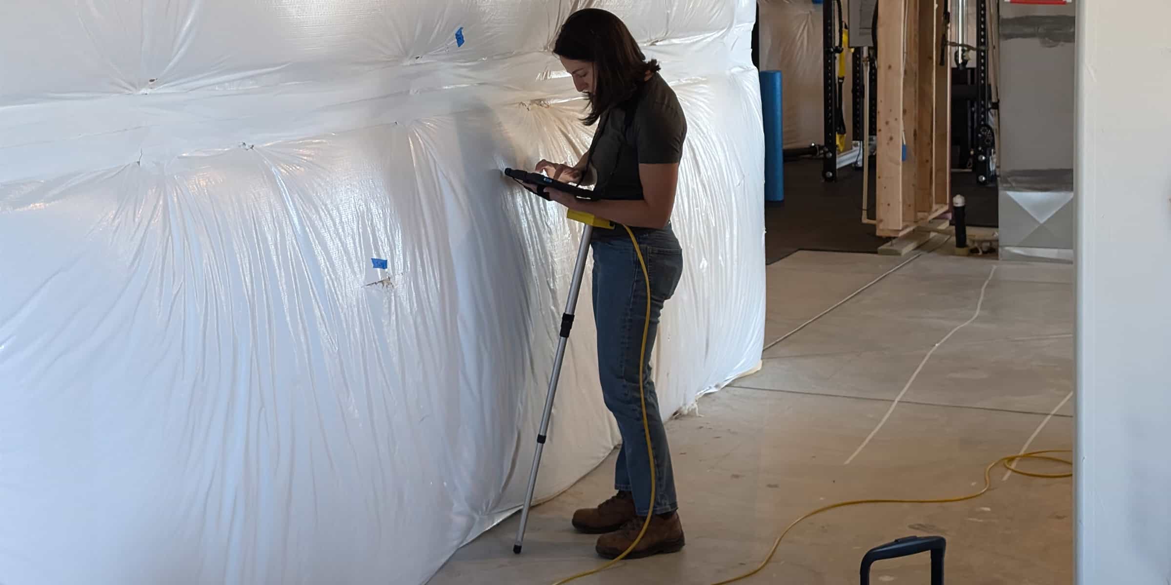 Woman using a tablet mounted on a surveying pole, tapping the screen while standing beside large plastic-wrapped insulation in a construction basement with exposed framing and concrete floor.