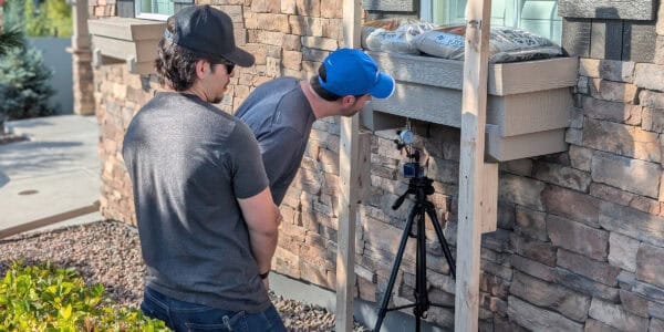 Two men examine a dial gauge on a tripod beneath a wooden window box attached to a stone house, leaning forward amid gravel and shrubs on a sunny front yard. 
Text visible: "PLAY SAND" (on bags atop the window box).