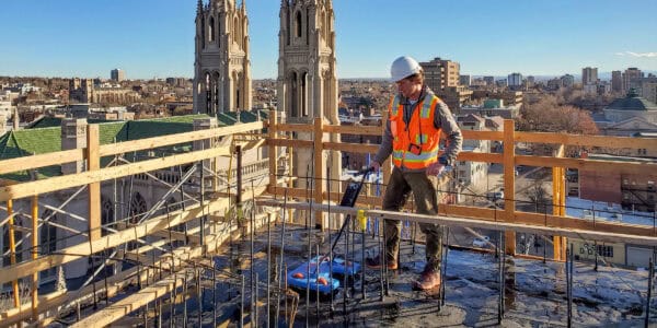 Construction worker wearing hard hat and orange safety vest operates a motorized concrete-smoothing device on a rooftop slab, surrounded by temporary wooden guardrails with cathedral towers and city skyline beyond.