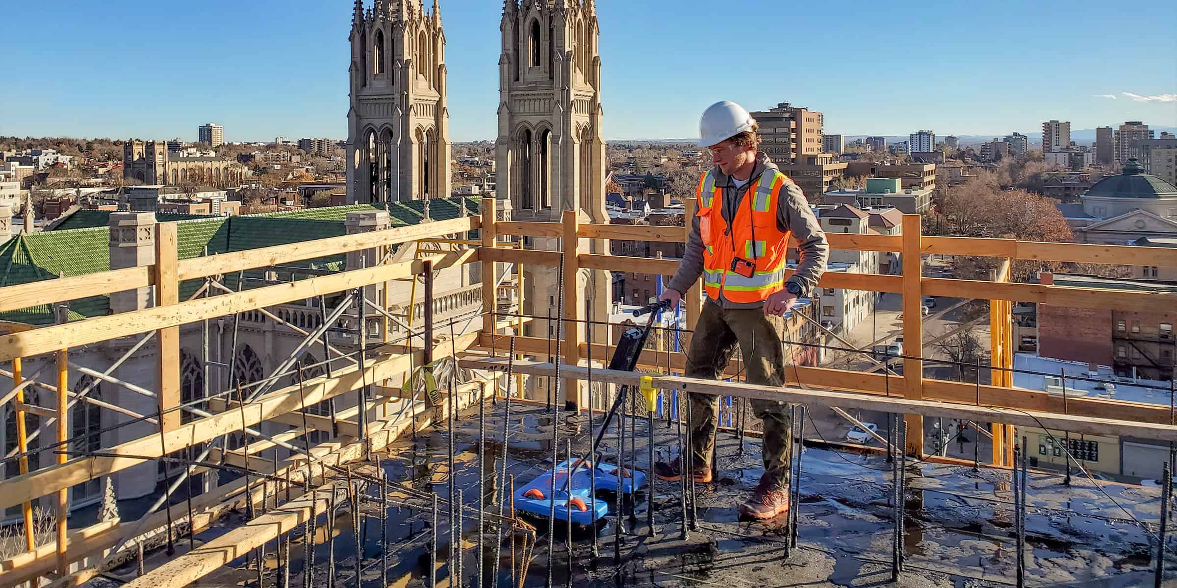 Construction worker wearing hard hat and orange safety vest operates a motorized concrete-smoothing device on a rooftop slab, surrounded by temporary wooden guardrails with cathedral towers and city skyline beyond.