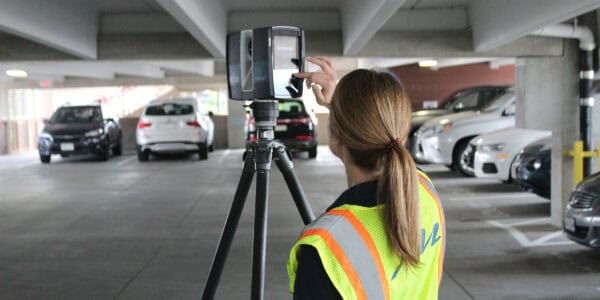 3D laser scanner on a tripod being touched by a technician adjusting its screen, in a dim multi-level parking garage among parked cars. Text visible: "FOCUS S" (scanner), "GITZO" (tripod), "ML" (vest).