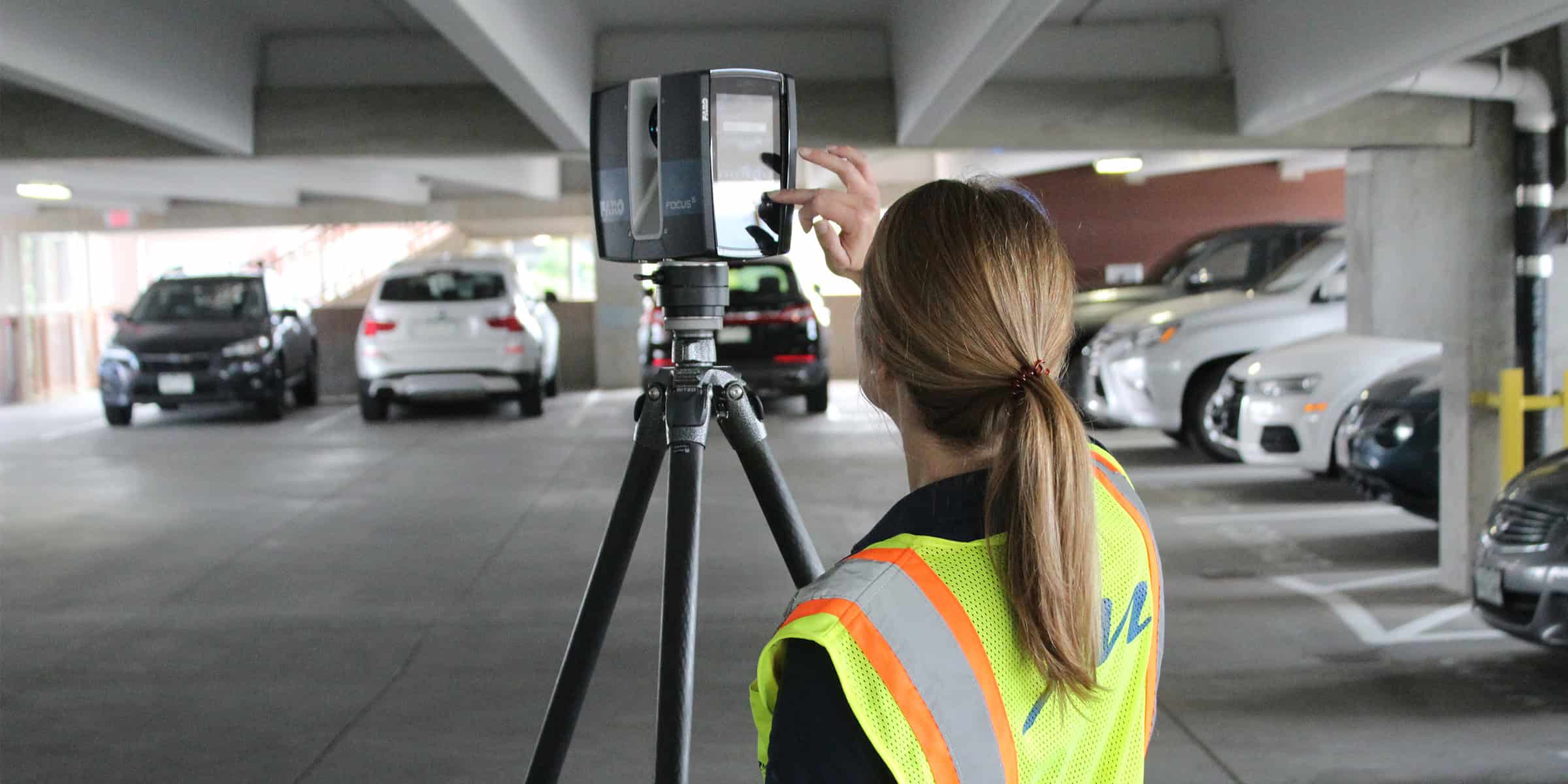 3D laser scanner on a tripod being touched by a technician adjusting its screen, in a dim multi-level parking garage among parked cars. Text visible: "FOCUS S" (scanner), "GITZO" (tripod), "ML" (vest).