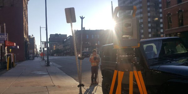 Survey tripod and total station stand in the foreground while a worker operates a rod-mounted device beside a parked vehicle on a sunlit downtown sidewalk and street.

Text found: "SP+" "Pay Here" "KING" "P"
