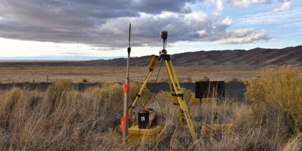 Survey tripod with GNSS receiver and marked pole collects data next to a battery case by a road amid dry grasses, open plain and rippled sand dunes beneath cloudy sky.