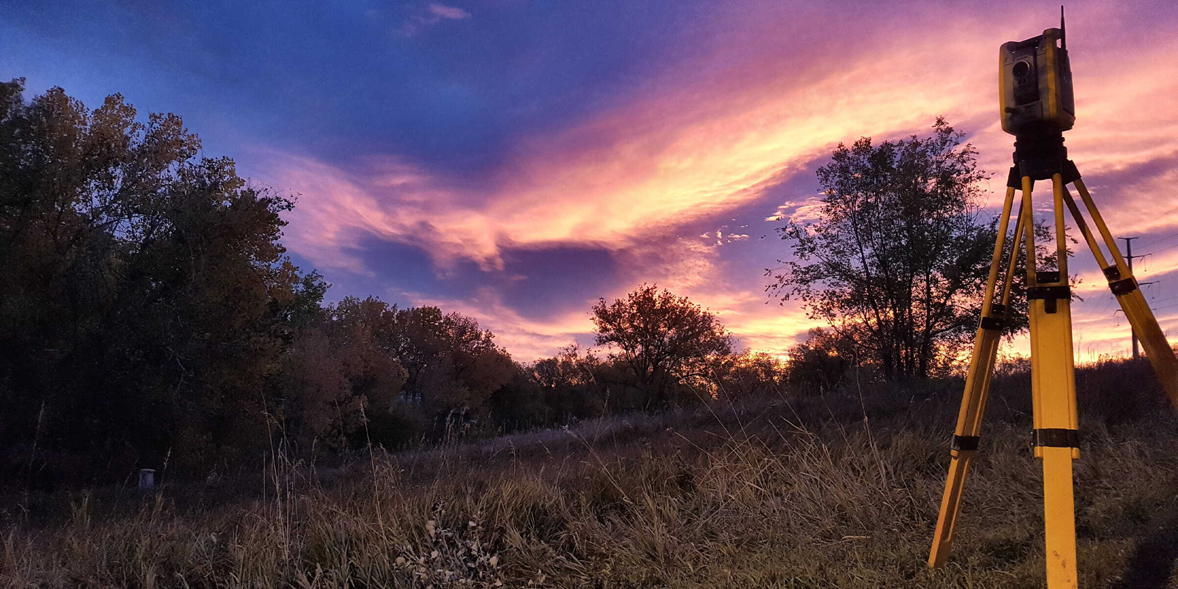 A yellow tripod-mounted surveying instrument stands overlooking a tree-lined field beneath a dramatic purple-and-pink sunset sky.