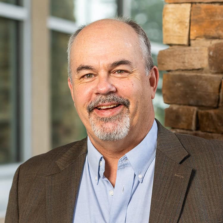 Middle-aged man with short graying hair and salt-and-pepper goatee smiles warmly at camera while wearing a brown blazer and light-blue button shirt, standing indoors by a stacked-stone column and window.