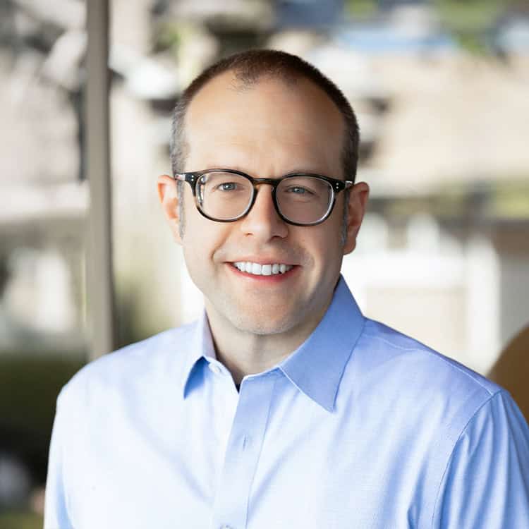 Adult man wearing glasses, smiling at the camera, in a light-blue button-up shirt, standing indoors near a window with a softly blurred exterior background.