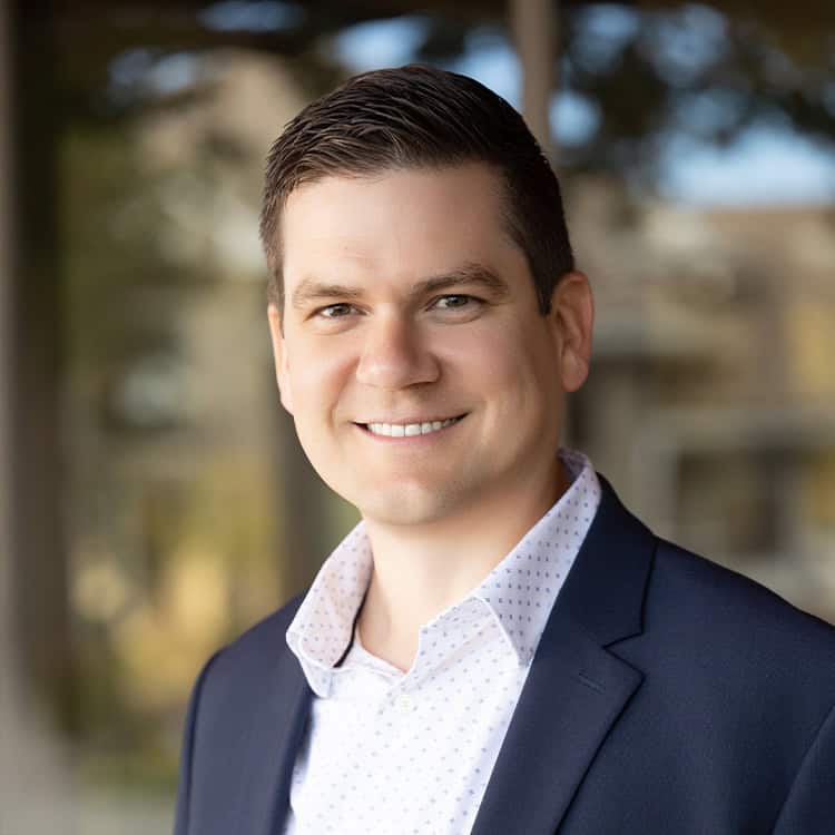 Smiling man in a navy blazer and patterned white shirt faces the camera, slightly angled; softly blurred outdoor urban background with glass reflections and trees.