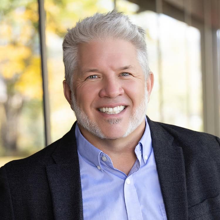 Smiling middle-aged person with white hair and trimmed beard, wearing a blue shirt and dark blazer, facing camera in a bright indoor setting beside a window with autumn foliage outside.