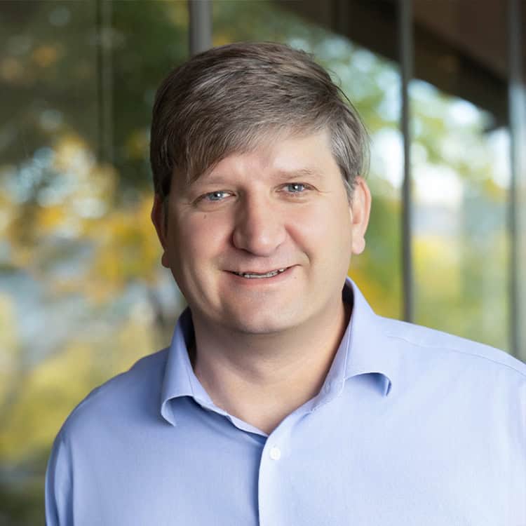 Man smiling, looking at the camera, wearing a light blue button-up shirt, standing indoors before large glass windows showing blurred green-and-yellow trees outside.