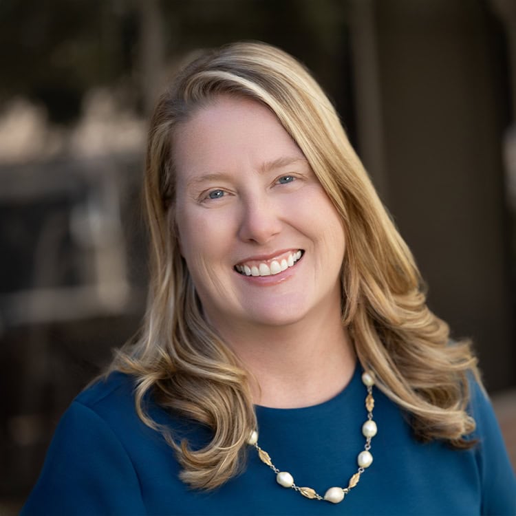 Woman smiling broadly, shows teeth, wearing blue top and pearl-and-gold necklace; shoulder-length blonde hair softly curled, posed against a softly blurred neutral outdoor background.