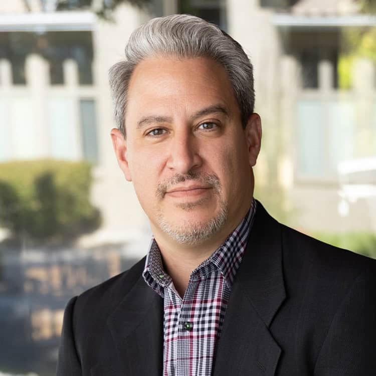 A middle-aged man with salt-and-pepper hair and trimmed goatee, looking at the camera with a slight smile; wearing a black blazer and checked shirt, blurred building and greenery behind.