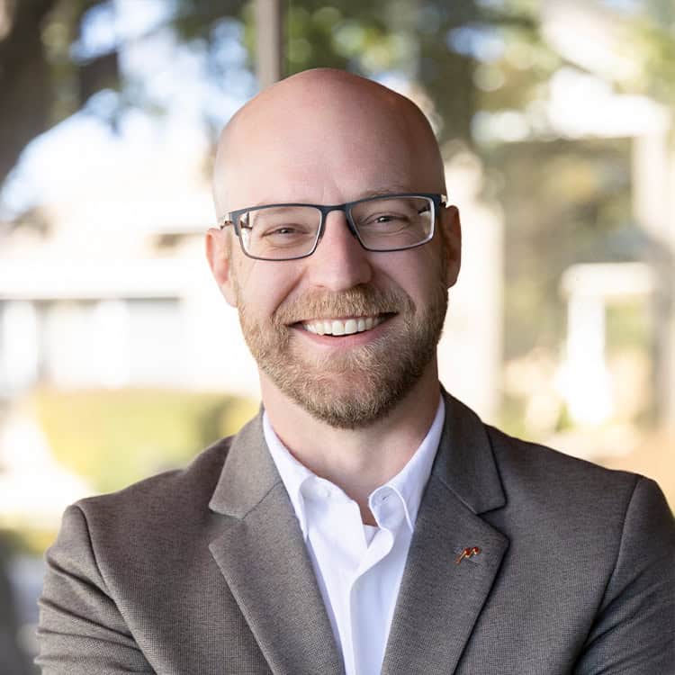 Bald man with glasses and trimmed beard smiles with arms crossed, wearing a gray blazer and white shirt, standing before a softly blurred outdoor/office background.