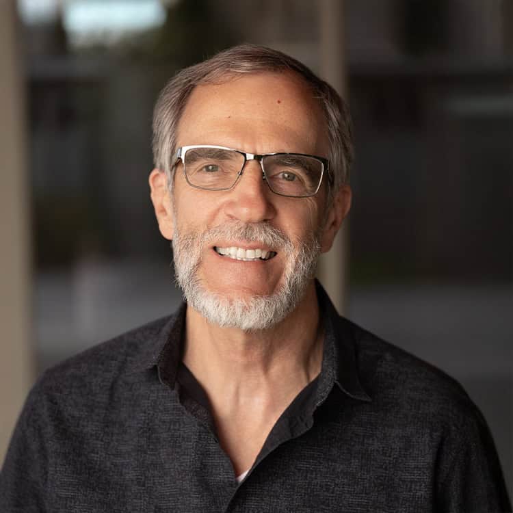 Older man with glasses smiles at the camera; white-gray beard and short hair, wearing a dark collared shirt against a softly blurred indoor background.