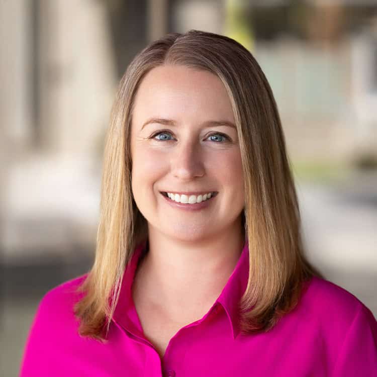 Woman smiling directly at camera, wearing a bright magenta blouse with shoulder-length straight blonde hair, against a softly blurred neutral background suggesting an indoor corridor or outdoor plaza.