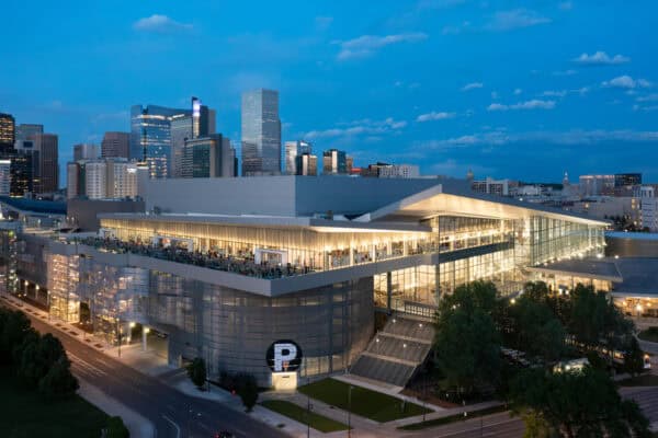 Glass-walled convention center glows with interior lights and a crowded rooftop terrace; people mingle while the downtown skyline rises behind at dusk, streets and trees framing the modern complex.

Visible text: "P"