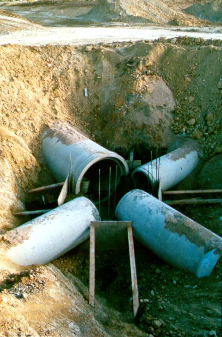 Four large concrete drainage pipes converge into a central junction, partially buried with exposed rebar and wooden braces, set within an excavated dirt pit at a construction site.